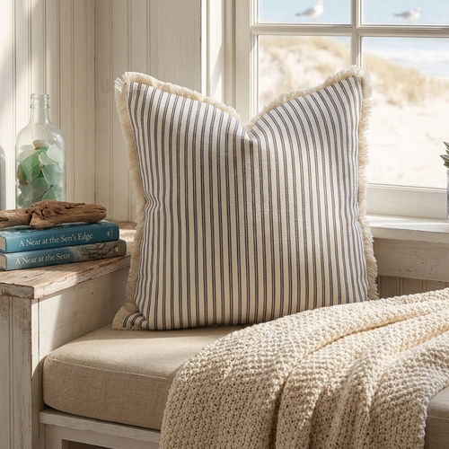 Striped pillow with fringe, on a cushioned bench with a window in the background