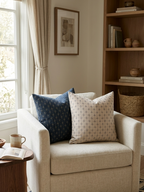 A cozy living room corner featuring a light beige armchair with two decorative throw pillows, one navy blue and one cream with a geometric pattern, set next to a wooden bookshelf. Pillow covers have a zipper to remove and wash covers. Inserts sold separately. 