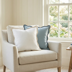 Beige armchair with white and blue washable throw pillows in a room with a window and floral view.