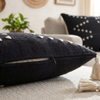 Black textured pillow with tassels on a neutral-colored surface, with another pillow and plant in the background.